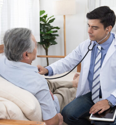 Caucasian doctor sit on chair check asian senior elderly old man Caucasian doctor sit on chair check asian senior elderly old man heart beat lay down on sofa at home.Medicine and healthcare concept.